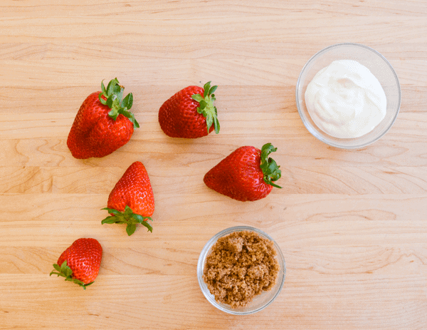 Fresh strawberries on a wooden cutting board with small bowls of brown sugar and sour cream.