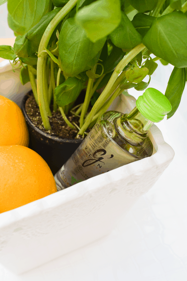 A mini bottle of gin in a white container with a basil plant and Meyer lemons to create a cocktail kit gift. 