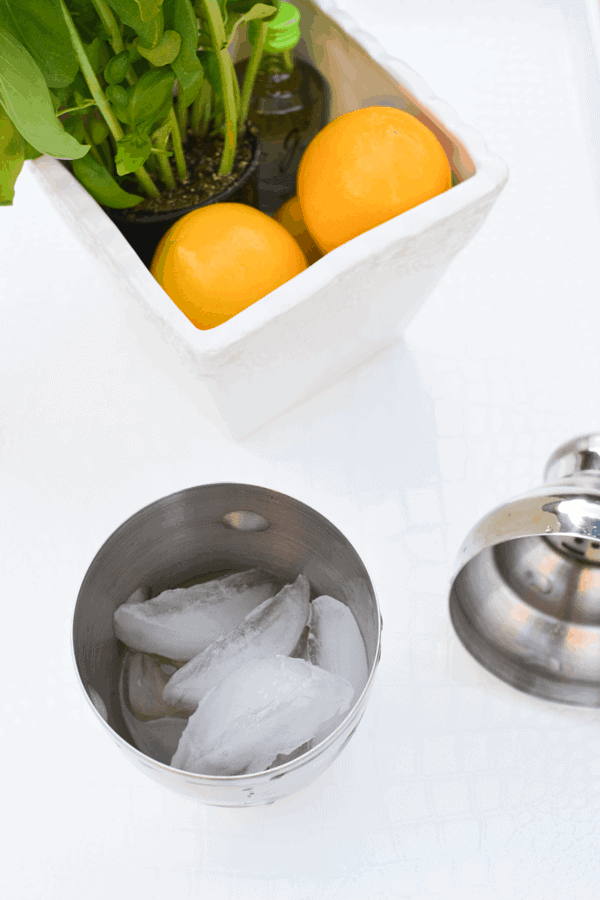 A cocktail shaker filled with ice on a table with the lid next to it and a container holding lemons and a basil plant.