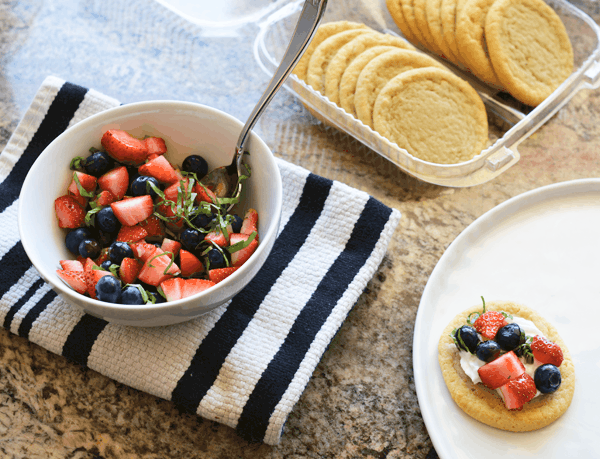 Cut up fresh berries and basil in a bowl sit next to a container of store-bought sugar cookies and a plate with a berry bruschetta waiting to be served.