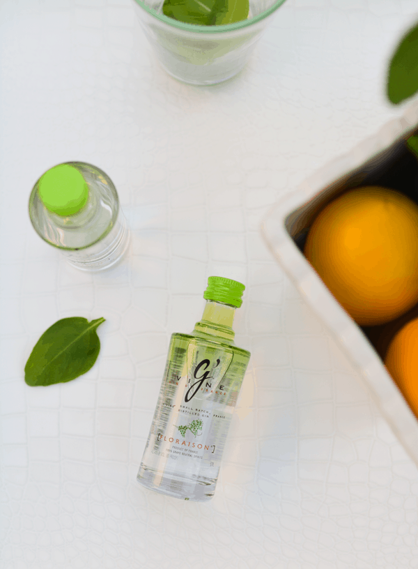 A small bottle of gin laying down on a table next to basil leaves and a white container holding lemons. 