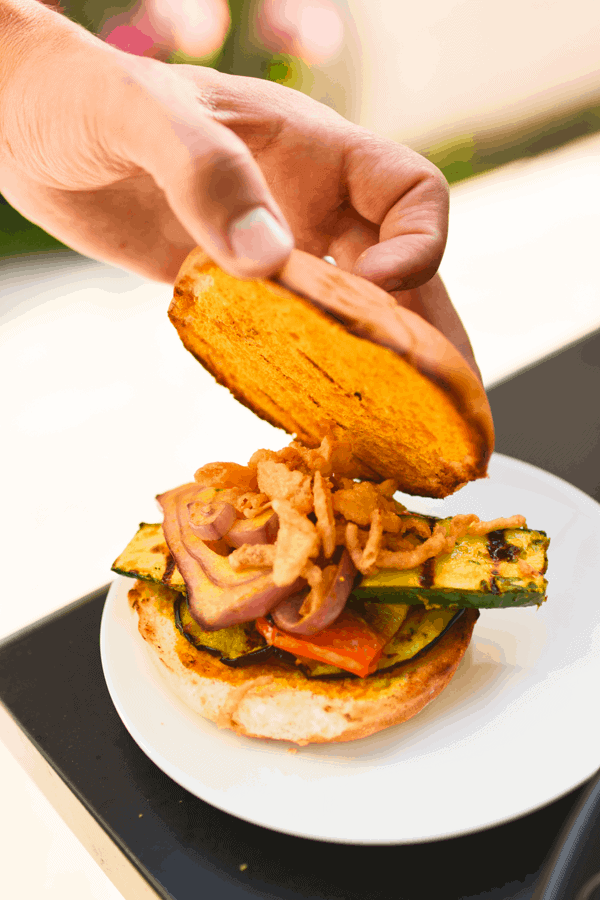 Grilled vegetable sandwich on a plate with someone adding a bun. 