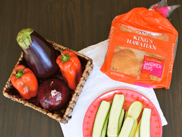 A package of King's Hawaiian Rolls on a table next to a basket of vegetables and a plate of sliced zuccini. 