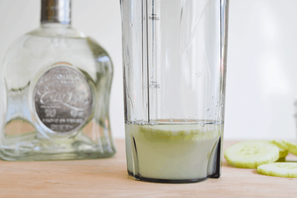 Close up of an individual sized plenter container with juice and cucumber in it with a bottle of tequila in the background.