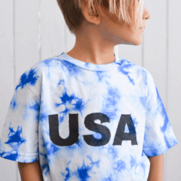 square photo of a boy in a blue and white tie dye shirt with the word USA spray painted on it.