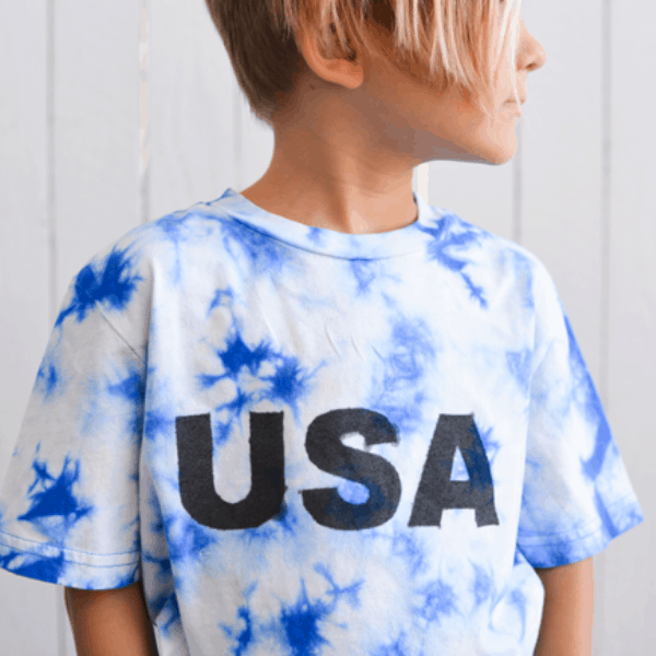 square photo of a boy in a blue and white tie dye shirt with the word USA spray painted on it.
