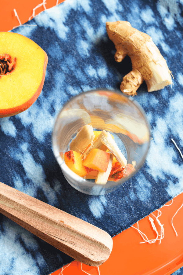 Peaches and ginger in the bottom of a glass next to a muddler and fresh peach and ginger pieces.