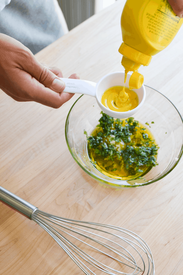 Adding yellow mustard to a measuring cup to add to a vinaigrette in a bowl on the counter. 