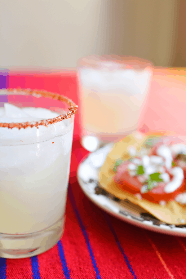 A cucumber margarita with Tajin on a table with a Tostada Caprese on a plate in the background.