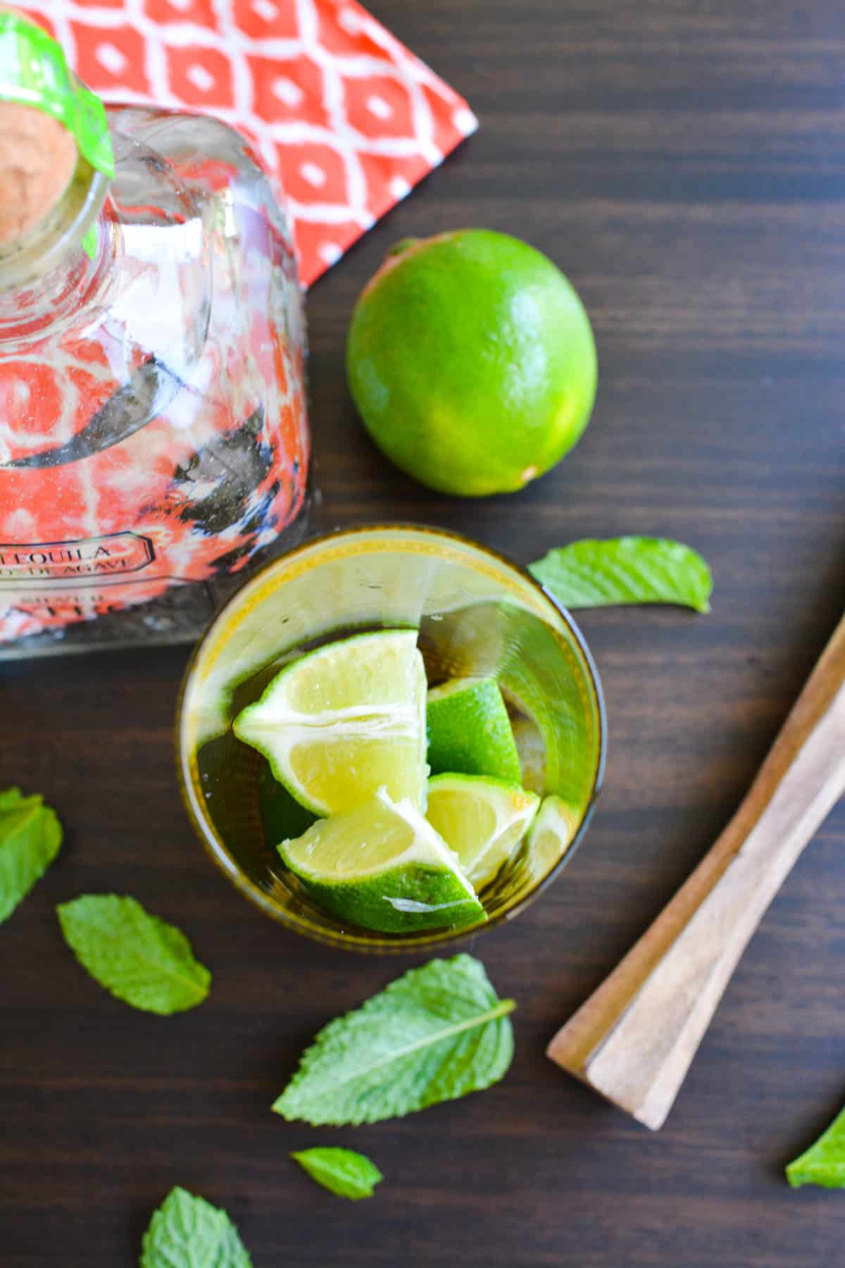 Overhead view of a glass with fresh lime pieces and mint next to a bottle of Patron tequila. 