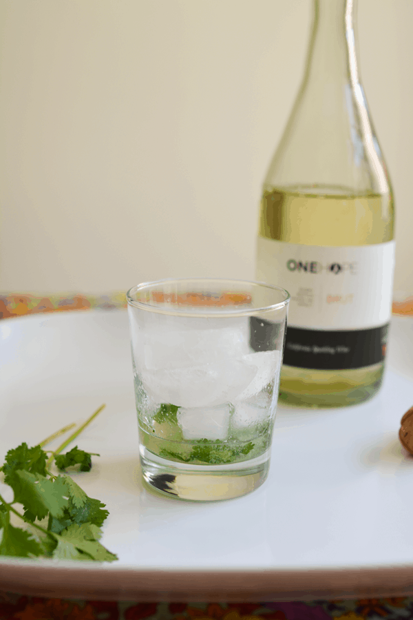 A cocktail glass full of ice with lime juice and cilantro on a white table next to a bottle of sparkling wine.