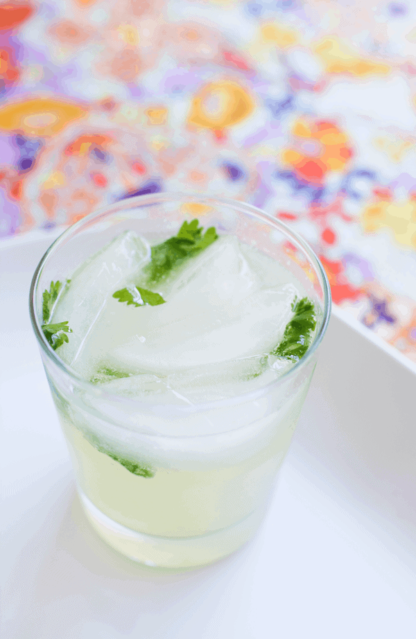 A cocktail glass with a drink, ice and cilantro on a white tray.