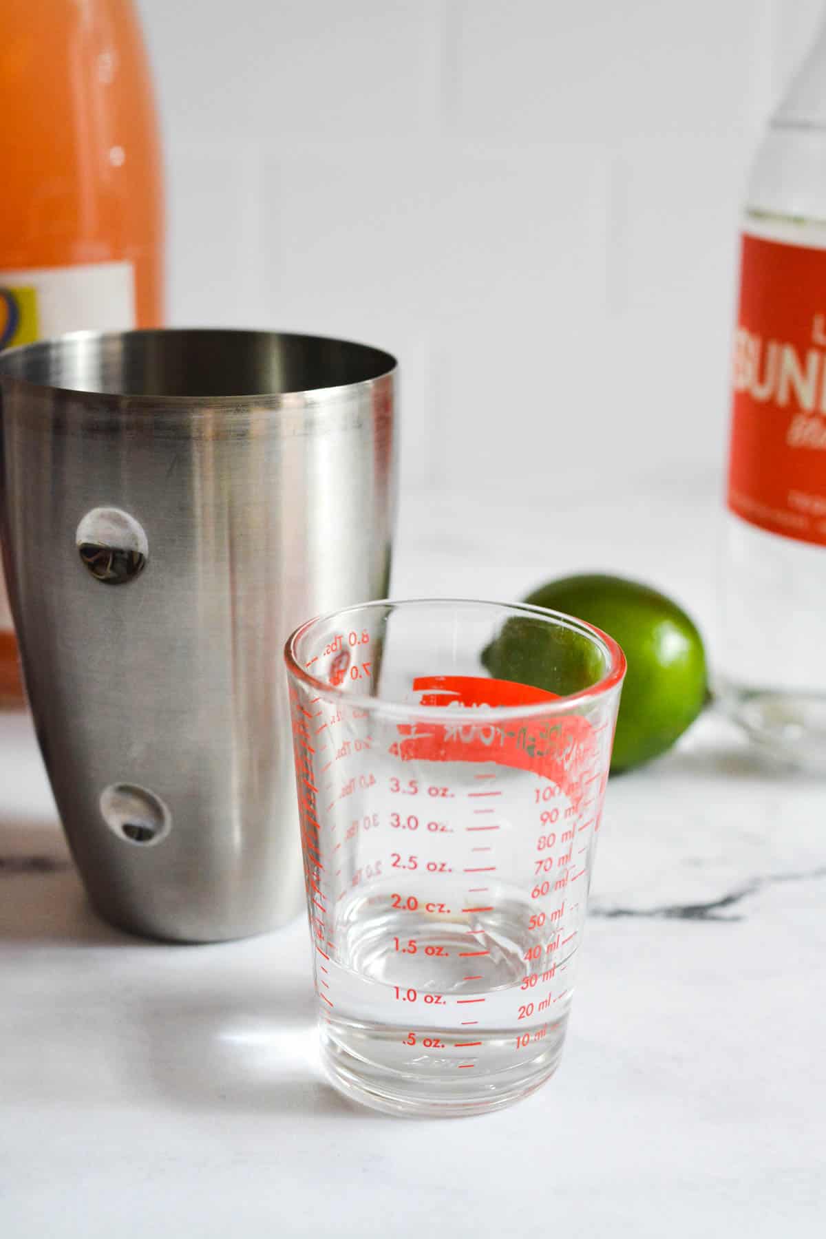 A measuring cup with tequila on a counter next to a cocktail shaker and lime.