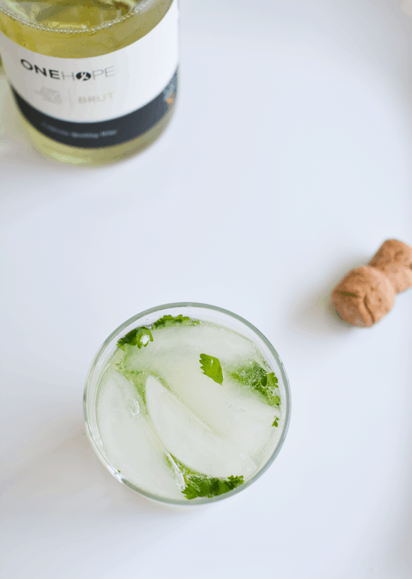 An overhead view of ice and cilantro in a cocktail in a glass on a white table next to a bottle of sparkling wine.