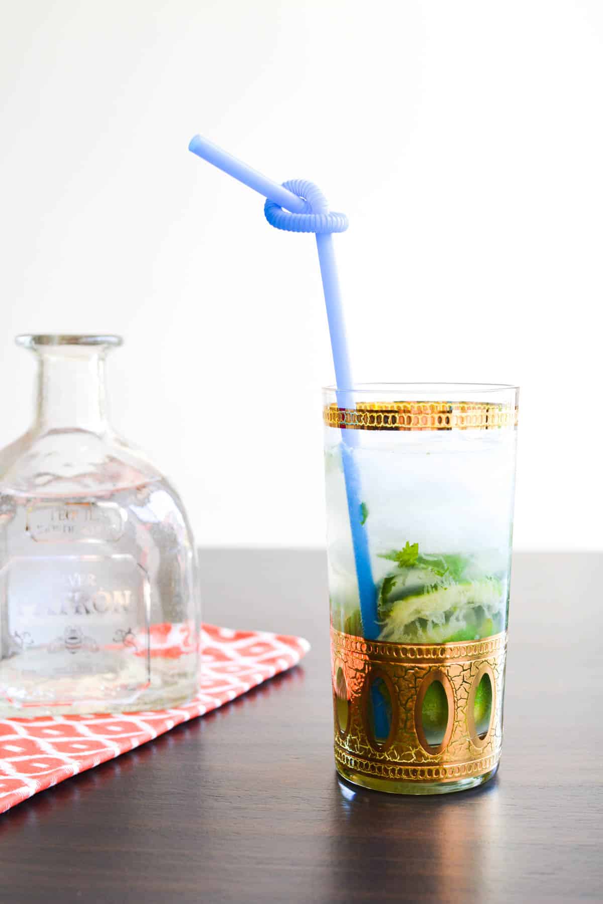 A cocktail glass with gold accents and a blue straw on a table next to a bottle of Patron tequila.