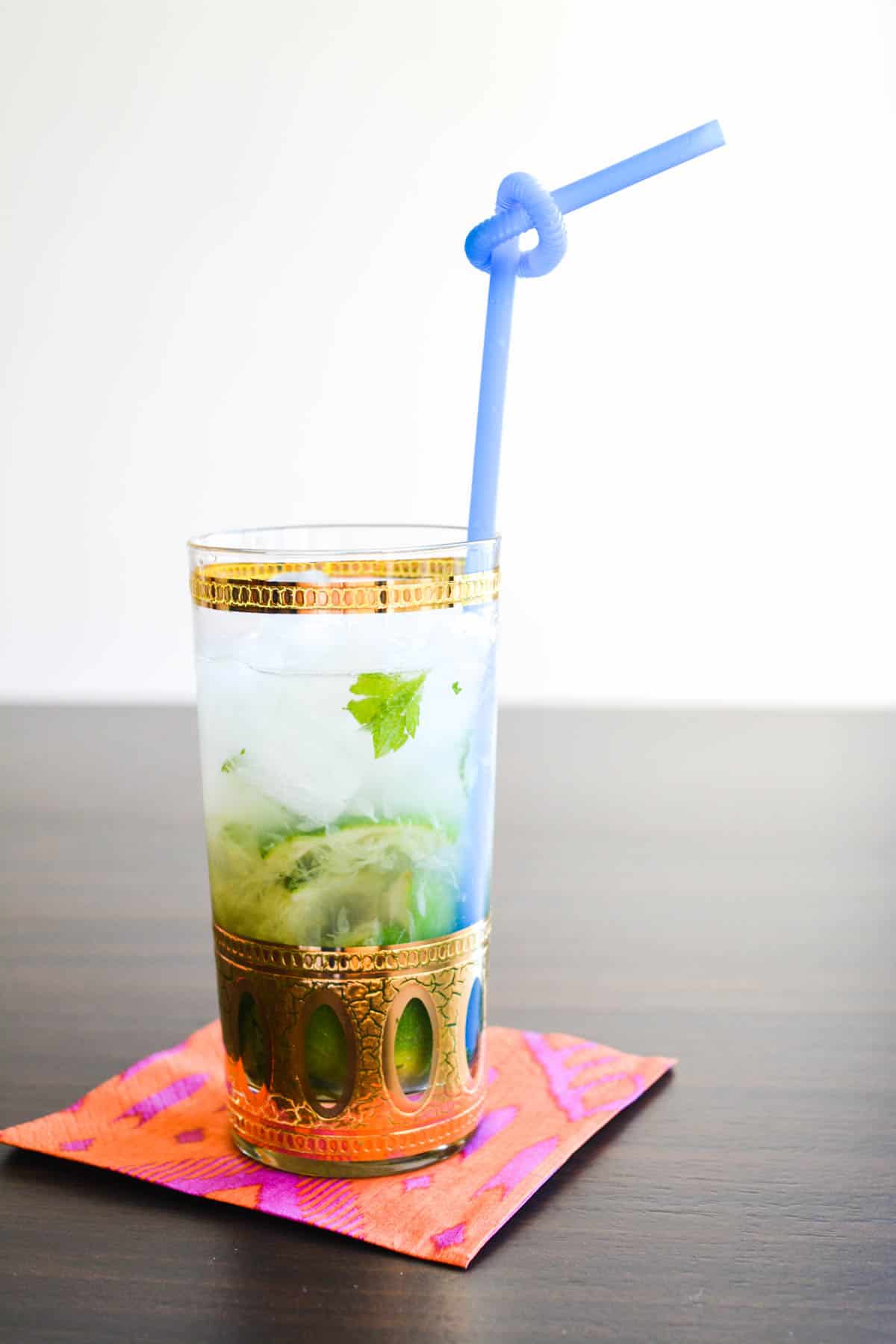 A cocktail glass on a napkin on a table with mint and lime in a clear liquid with blue bendy straw.
