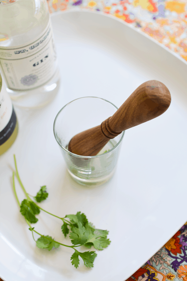 A cocktail glass on a white table with a wooden muddler and  cilantro sprigs next to it.