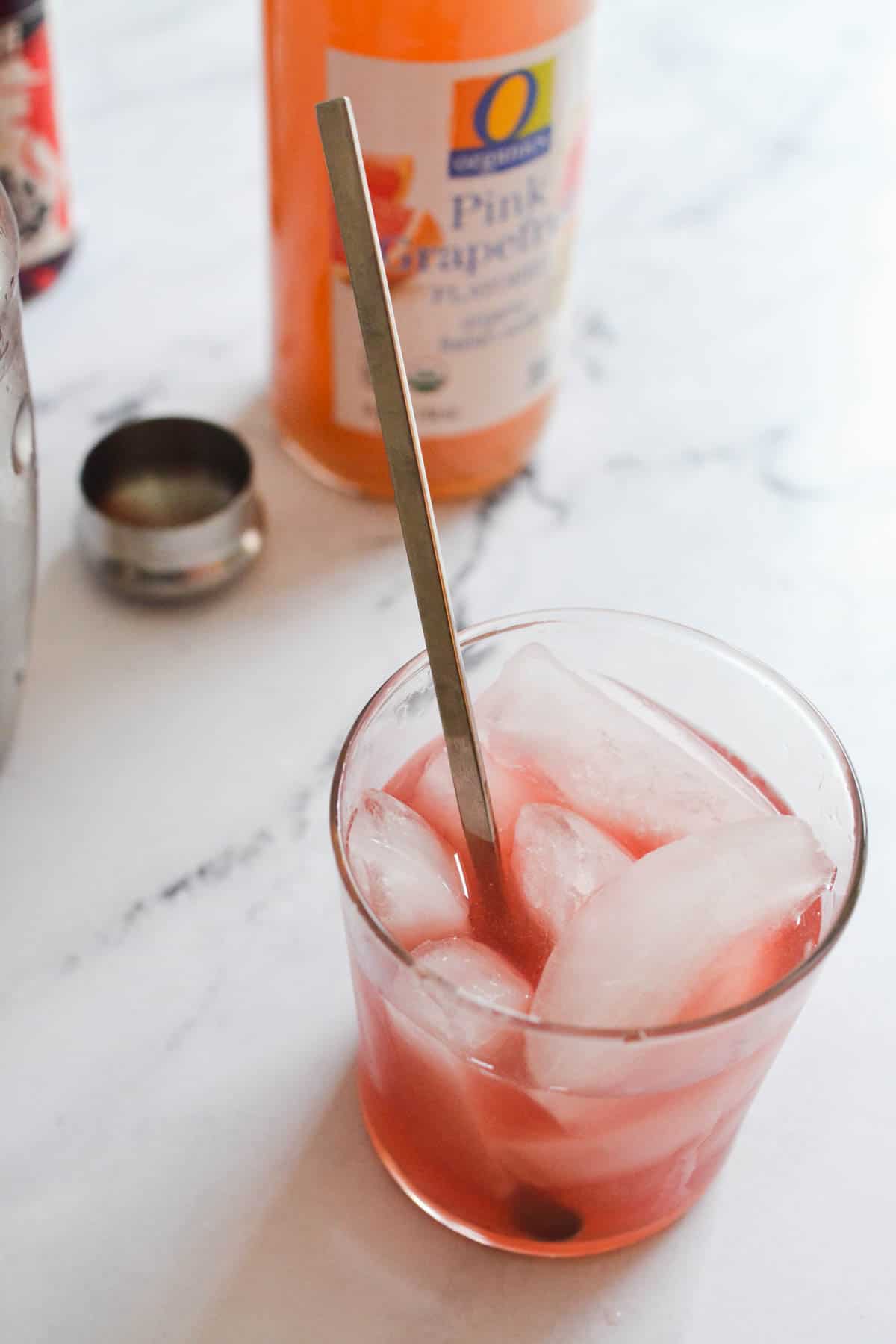 A cocktail on a counter with a spoon in it next to a bottle of grapefruit soda.