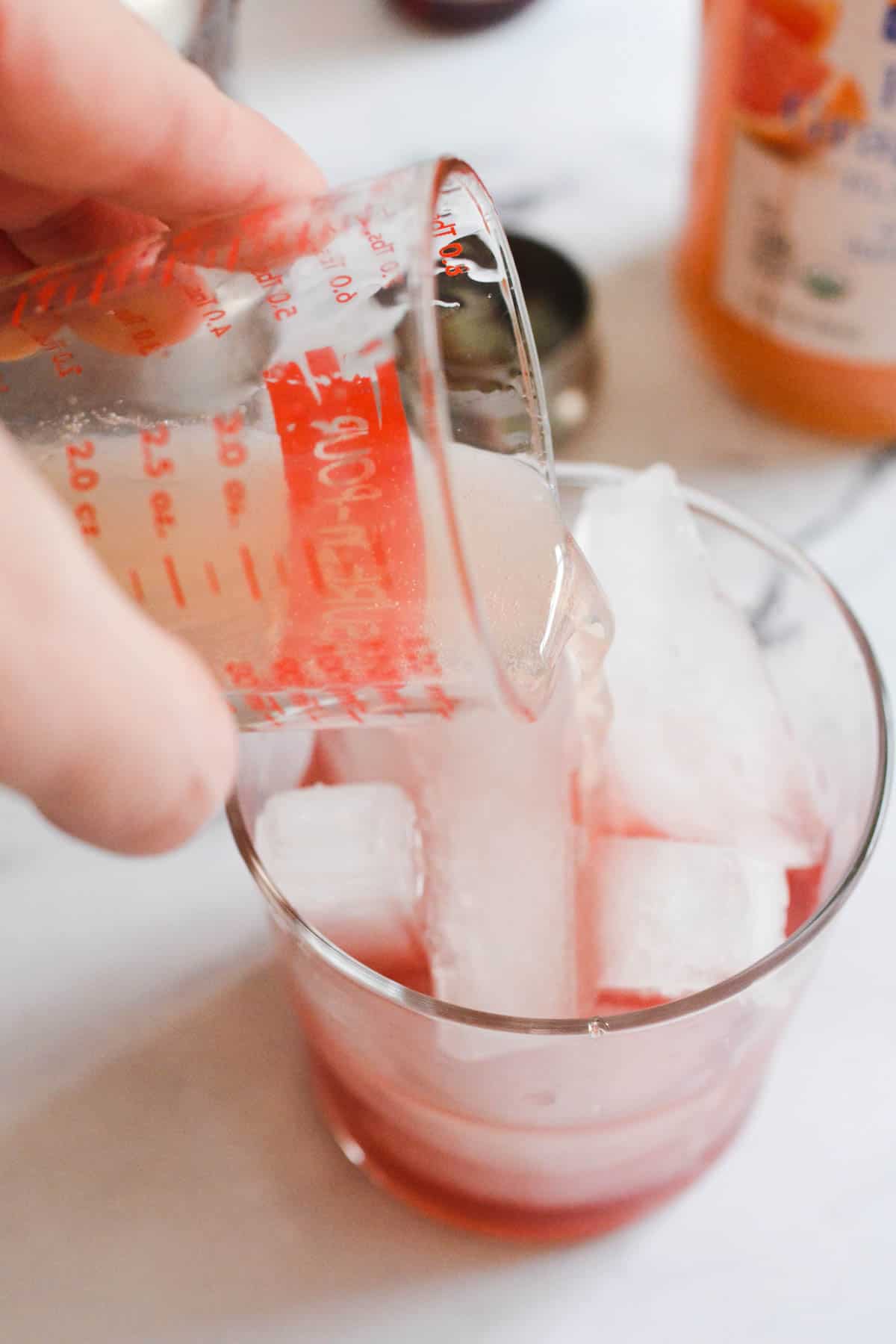 Close up of a measuring cup pouring grapefruit soda into a cocktail with ice.