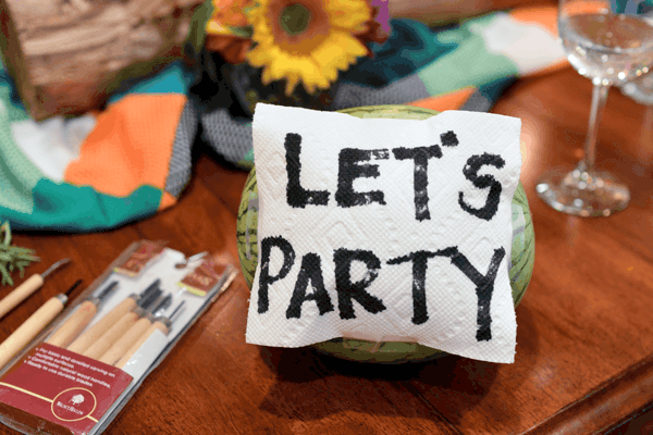 A watermelon with a napkin taped to it with a drawn message that says "Let's Party".