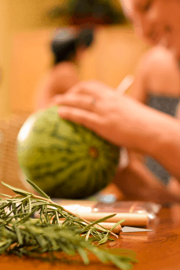 Close up of some fresh rosemary with a woman carving a watermelon in the background. 