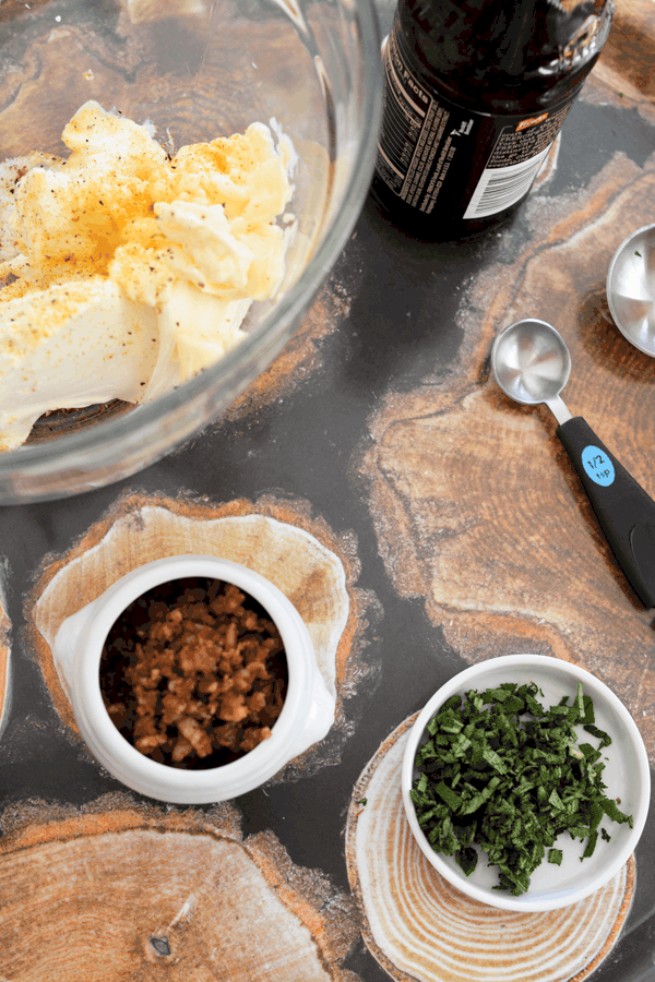 A small bowl of bacon bits, a dish of chopped fresh sage on a tray next to a mixing bowl with dip ingredients.