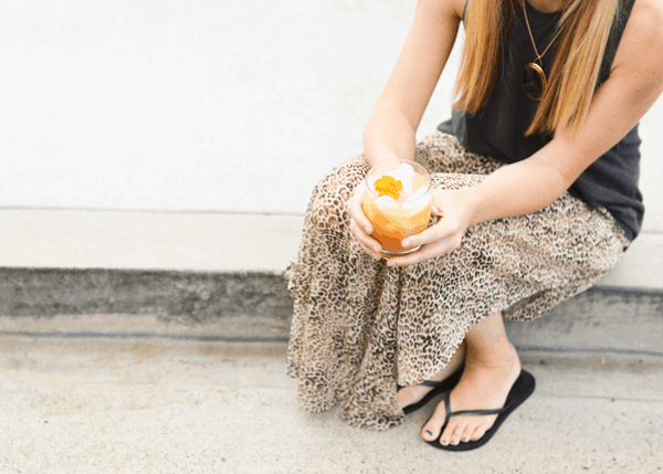 Girl sitting on a curb holding a Pear cocktail topped with an edible flower. 