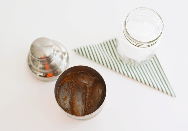 A jar full of ice on a napkin on a table next to an open cocktail shaker with iced coffee in it. 