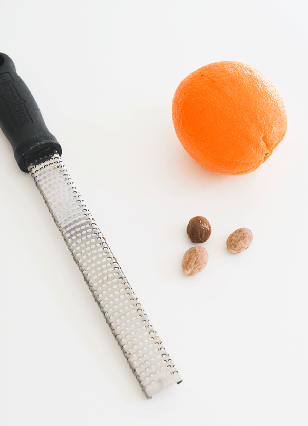 A Microplane zester on a table next to an orange and whole nutmeg.