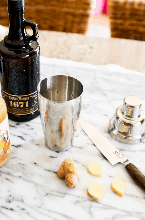 A cutting board with sliced ginger next to a knife and cocktail shaker and a bottle of spiced rum next to it. 