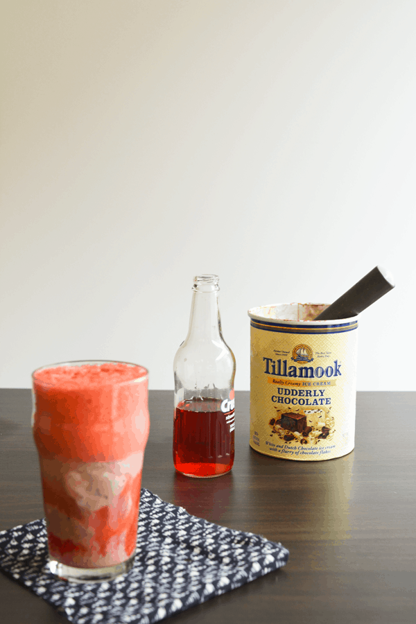 An ice cream float in a glass on top of a black and white napkin, in front of a bottle of soda and a carton of ice cream with a scoop inside. 