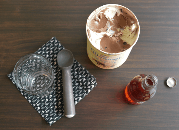 An open carton of ice cream next to an open bottle of soda and an ice cream scoop and a glass on top of a napkin, all on top of a wood surface.
