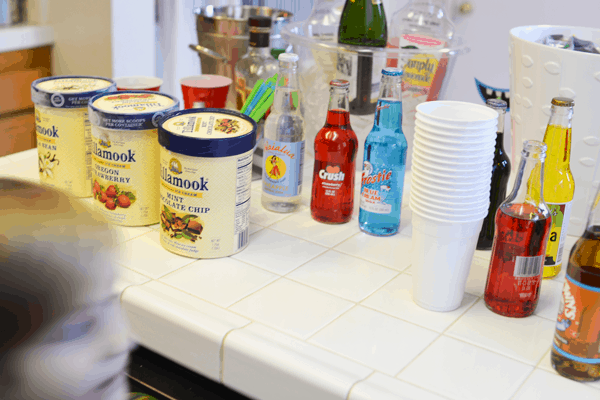 A countertop covered in flavored sodas, ice cream and cups for a drink station.