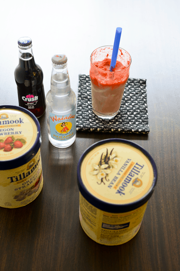 Cartons of ice cream and bottles of flavored soda in front of a glass filled with an ice cream float and a blue straw on a wood table.