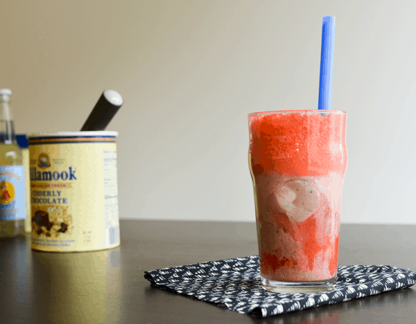 A strawberry soda and chocolate ice cream float on top of a black and white napkin in front of ice cream and a bottle of soda.