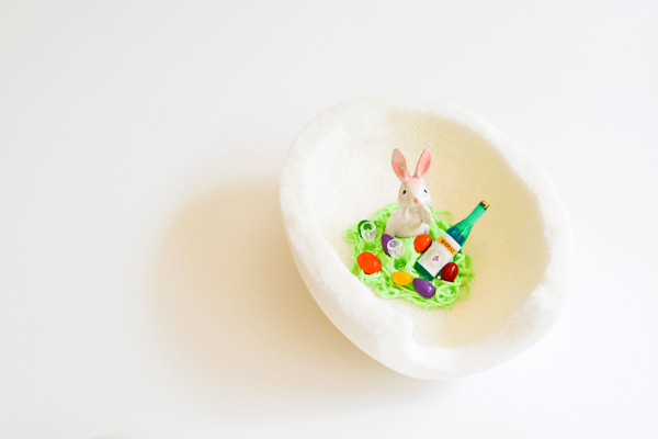 Half of a white sugar Easter Egg with a bunny miniature in frosting grass with mini Easter eggs and miniature of a bottle of wine.