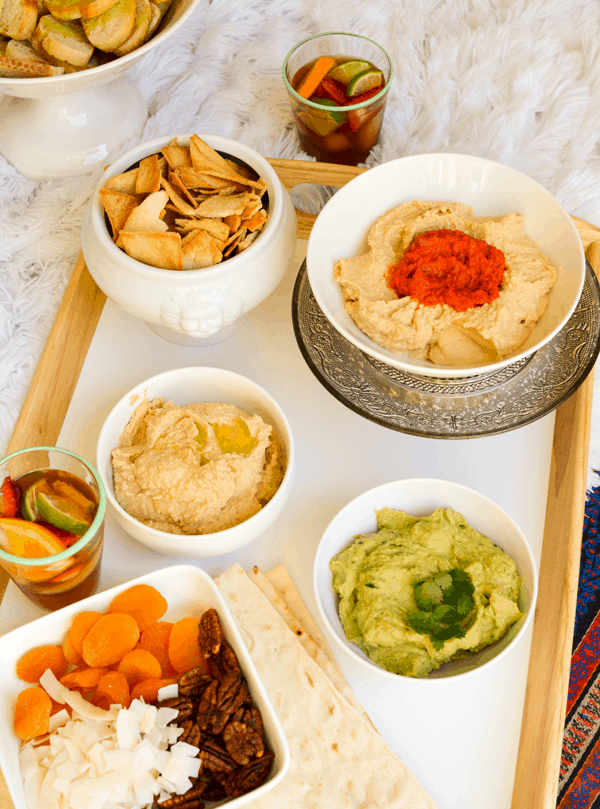 An overhead shot of a tray of food with white bowls of hummus, avocado hummus and chips. 