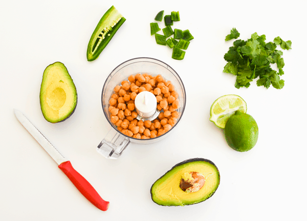 A small food processor with chickpeas in it next to cilantro, lime, avocado and jalapeno.
