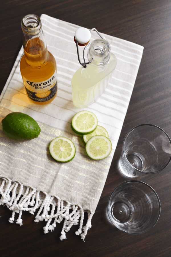 A striped towel beneath a corona, limeade and fresh limes to make a corona chelada.