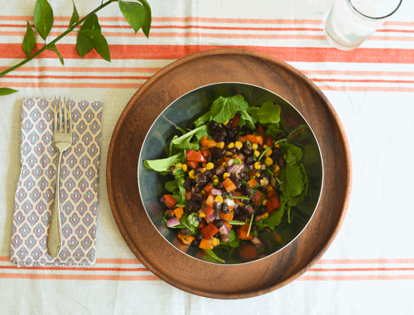 A bowl holding a Black Bean Salad over arugula on a table next to a fork.