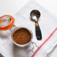 Glass jar filled with seasoning on a towel next to a measuring spoon.