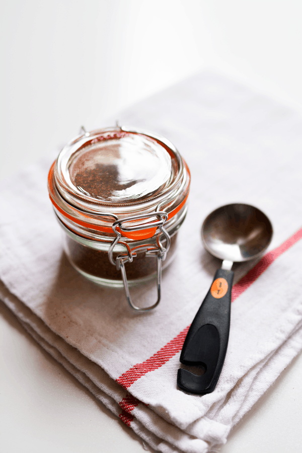 Small glass canister with seasonings inside on a towel next to a measuring spoon. 