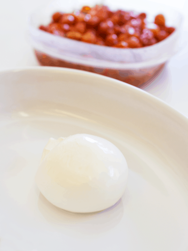 A ball of burrata cheese in a serving dish next to a container of roasted cherry tomatoes. 