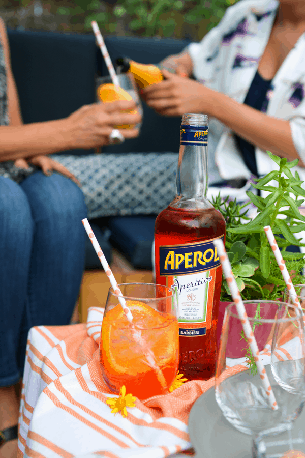 An Aperol Spritz sitting in a glass on a table next to a bottle of Aperol with two girl's cheersing their cocktails in the background.