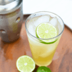 Spicy Serrano Margarita garnished with lime and ice on a wooden board next to a silver cocktail shaker.