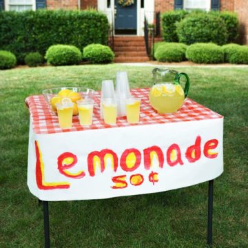 A lemonade stand on a lawn in front of a house.