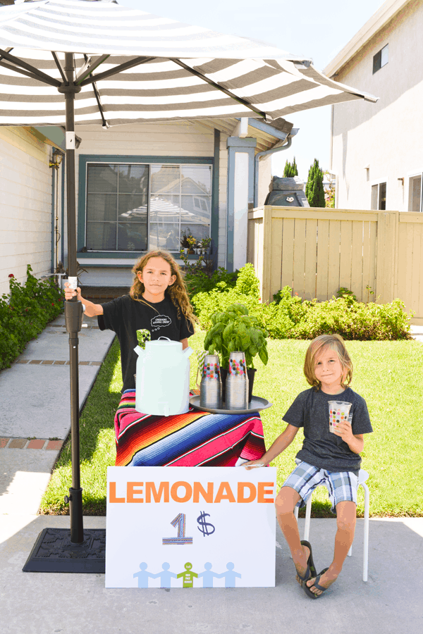 Two kids standing next to a lemonade stand in front of a house.