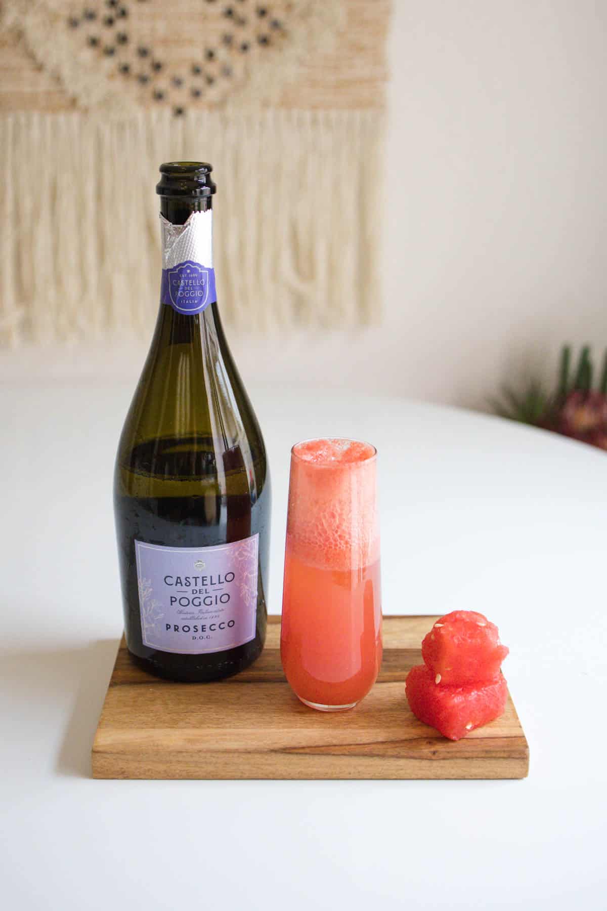 A watermelon mimosa on a wood tray next to a bottle of sparkling wine and pieces of watermelon.