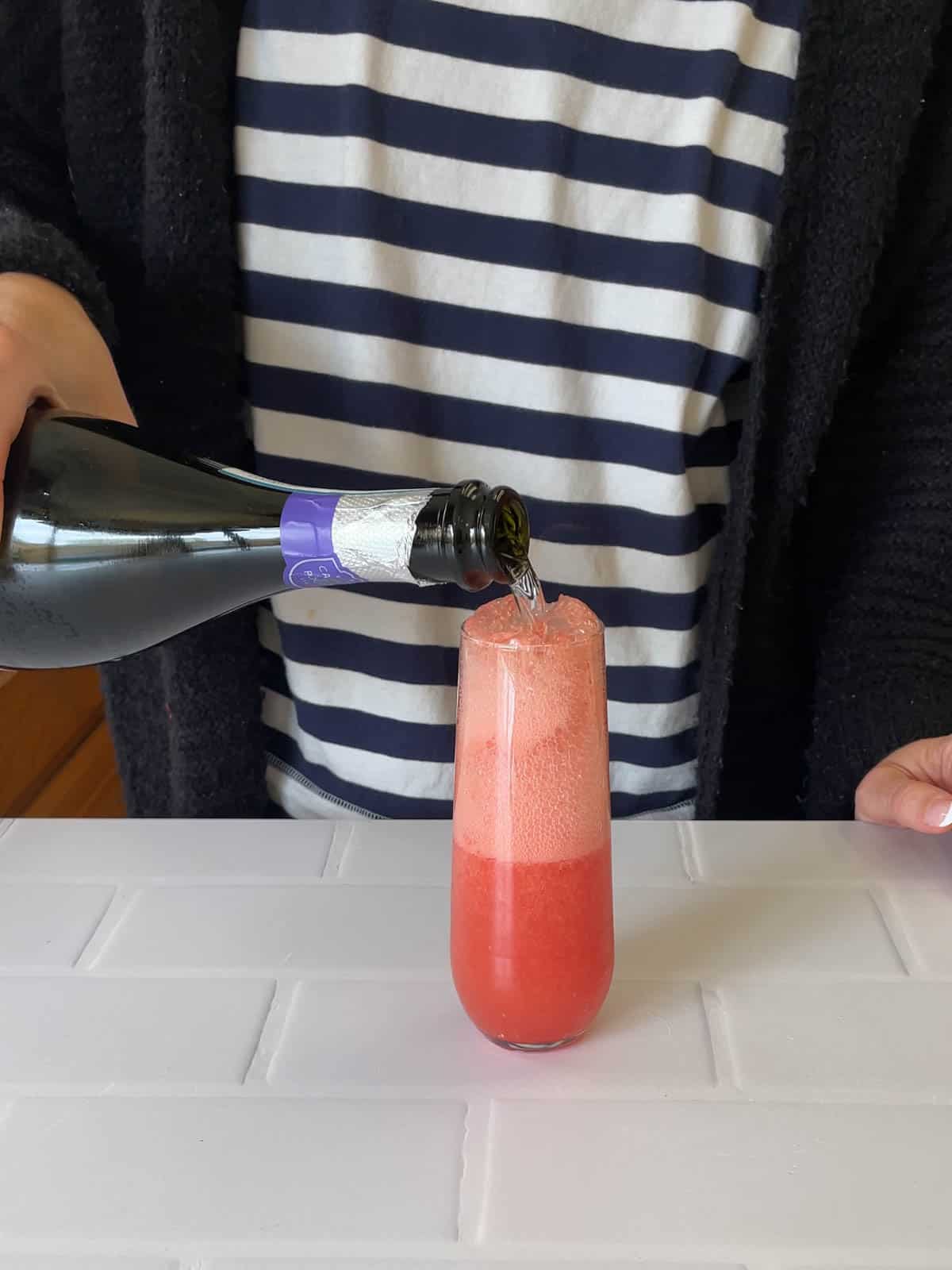 A woman adding champagne to a watermelon mimosa in a stemless flute.