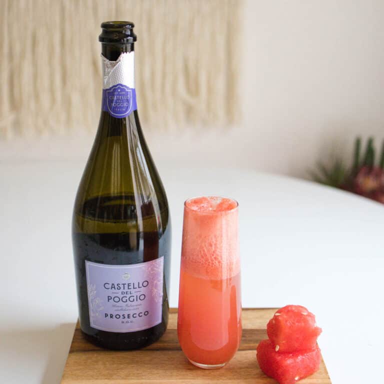 A bottle of champagne, a watermelon mimosa and 2 pieces of watermelon on a wood tray.